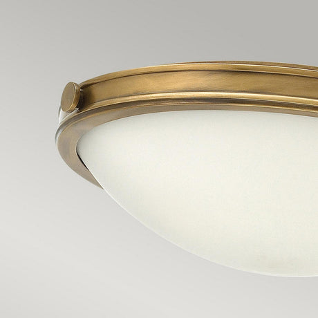 A ceiling-mounted Collier 3 Light Large Flush fixture features a dome shape with a Heritage Brass frame and frosted glass diffuser, presented against a plain gray background. The design is highlighted by a decorative circular accent along the edge, evoking the style of vintage glass shades.