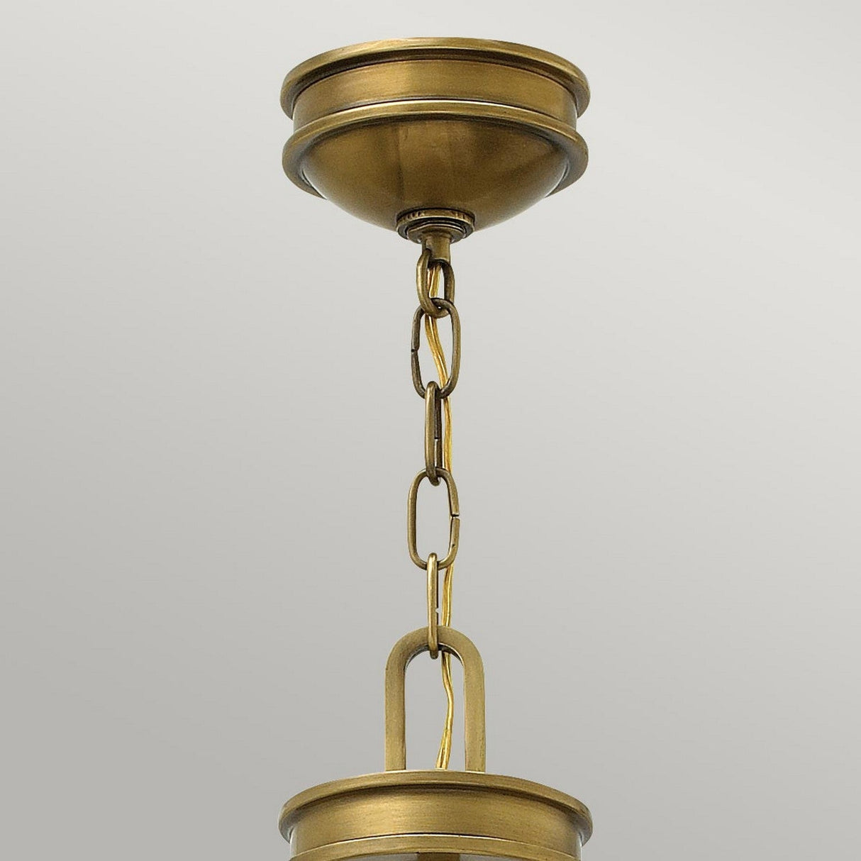A close-up of the Collier 4 Light Chandelier in Heritage Brass showcases a ceiling light fixture with a chain connecting to its dome-shaped base. The chandelier's simple, elegant design features smooth curves and links, exuding mid-century charm against a plain grey background.