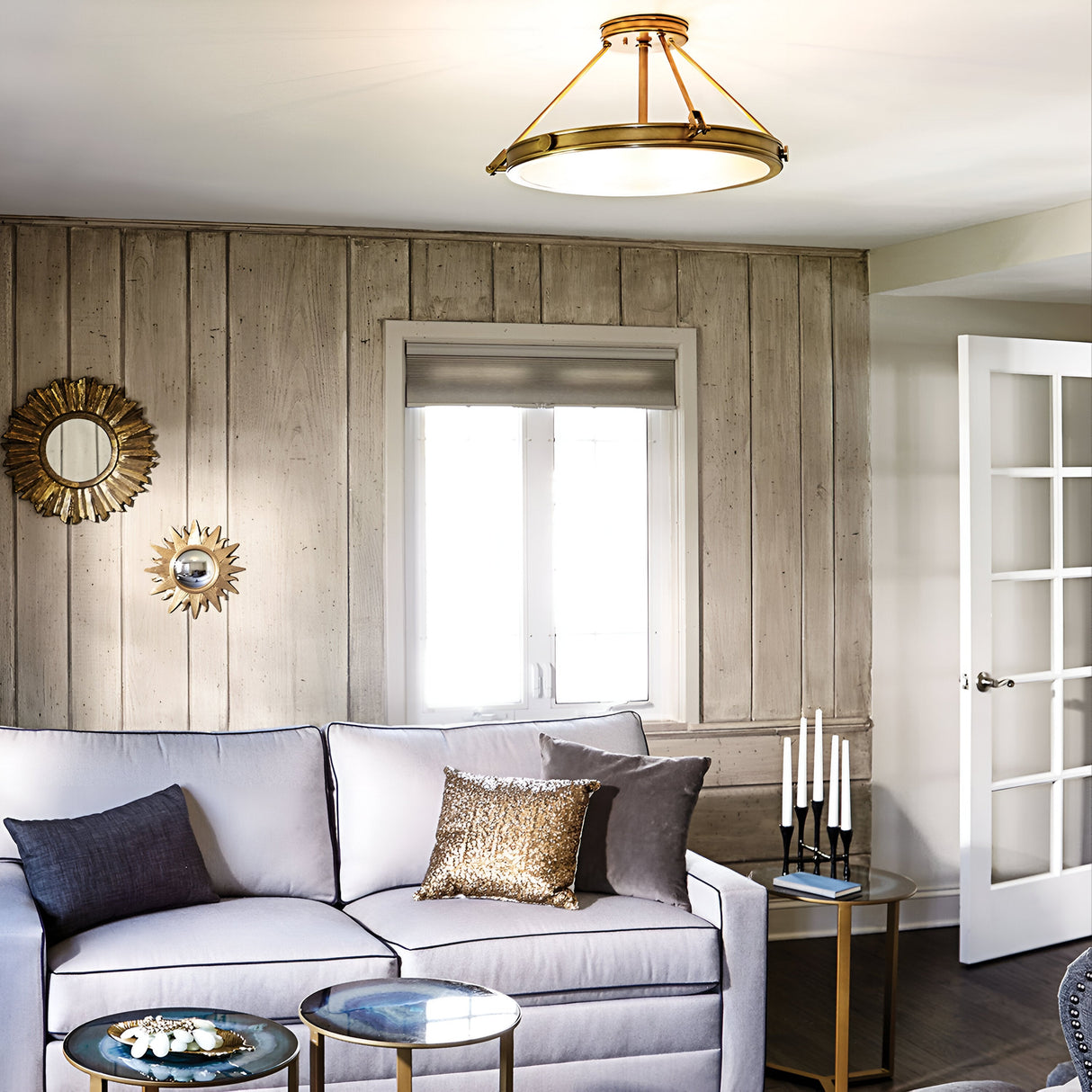 A cozy living room with light wood panel walls features a gray sectional sofa adorned with dark and gold pillows. A Collier 4 Light Large Semi Flush in Heritage Brass elegantly hangs from the ceiling. Two sunburst mirrors are on the wall, complemented by a white French door and side table with candles.