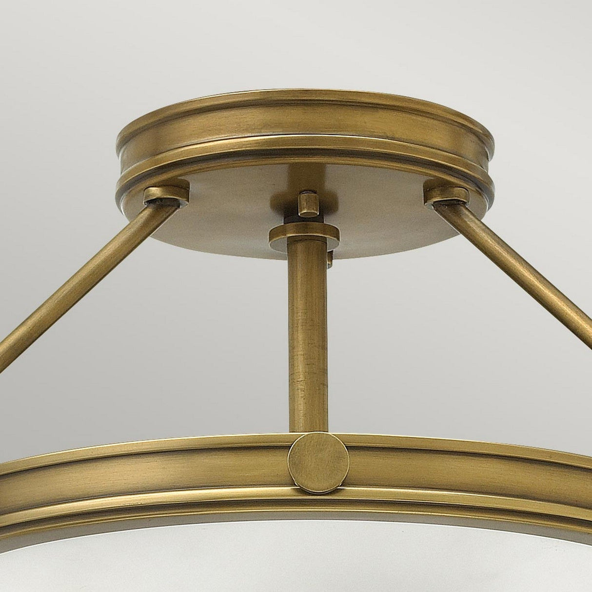 A close-up of the Collier 3 Light Semi Flush fixture in Heritage Brass showcases its circular design. With a sleek metallic finish and support from three angled rods, it presents an elegant and modern look against a simple backdrop.
