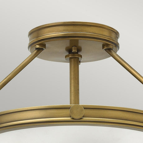 A close-up of the Collier 3 Light Semi Flush fixture in Heritage Brass showcases its circular design. With a sleek metallic finish and support from three angled rods, it presents an elegant and modern look against a simple backdrop.