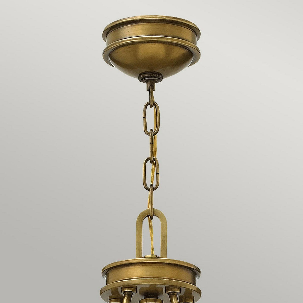 A close-up of the Collier 5 Light Chandelier in Heritage Brass showcases its linked chain detail. The fixture's canopy is mounted on a light gray ceiling, with a series of chain links leading to a decorative brass component below that evokes a Mid-Century silhouette.