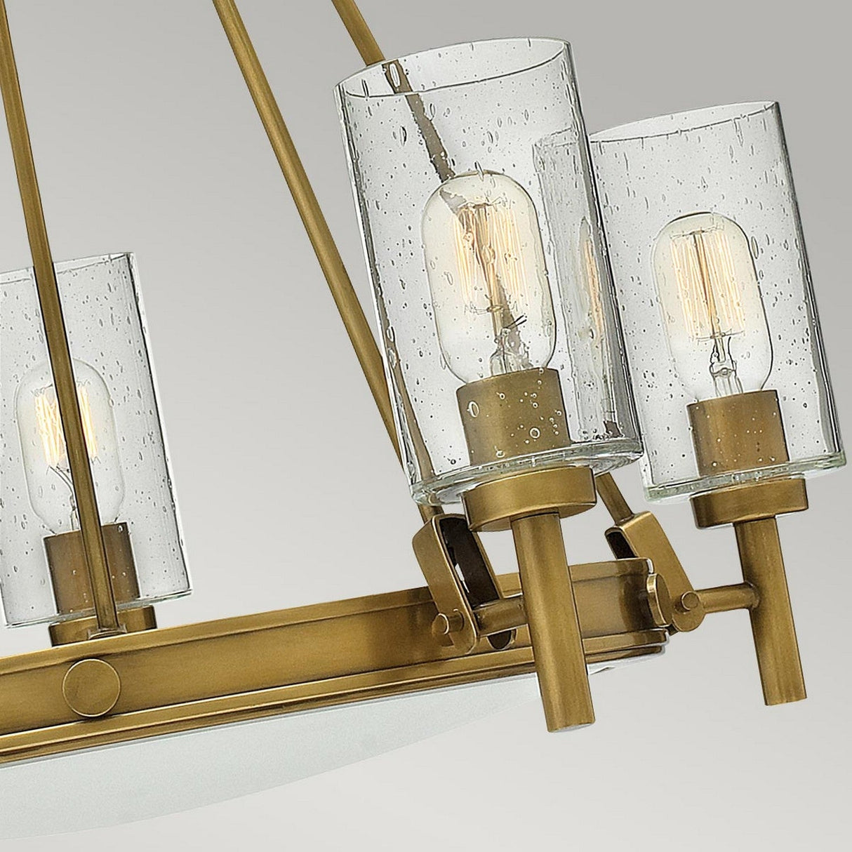 A close-up of the Collier 5 Light Chandelier - Heritage Brass, reveals each shade featuring a visible bulb encased in speckled glass. This elegant piece blends a mid-century silhouette with a modern, minimalist design set against a neutral backdrop.