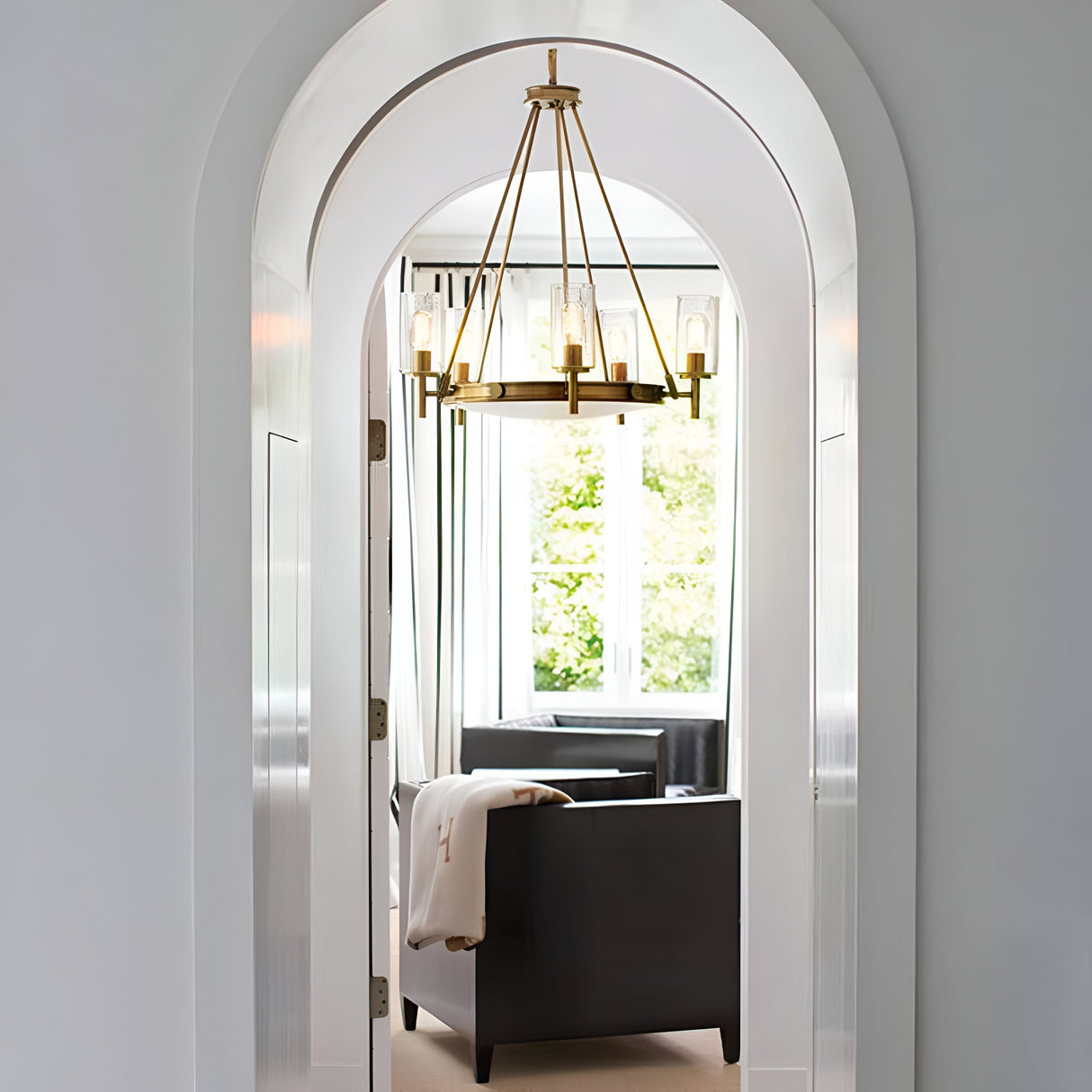 You can see through an arched doorway into a luminous room featuring a large window. Within the space, there's a black couch adorned with a cream throw and an elegantly sleek mid-century silhouette chair. Suspended from the ceiling is the Collier 5 Light Chandelier in Heritage Brass, enhancing the room with its modern design as soft natural light fills the area.