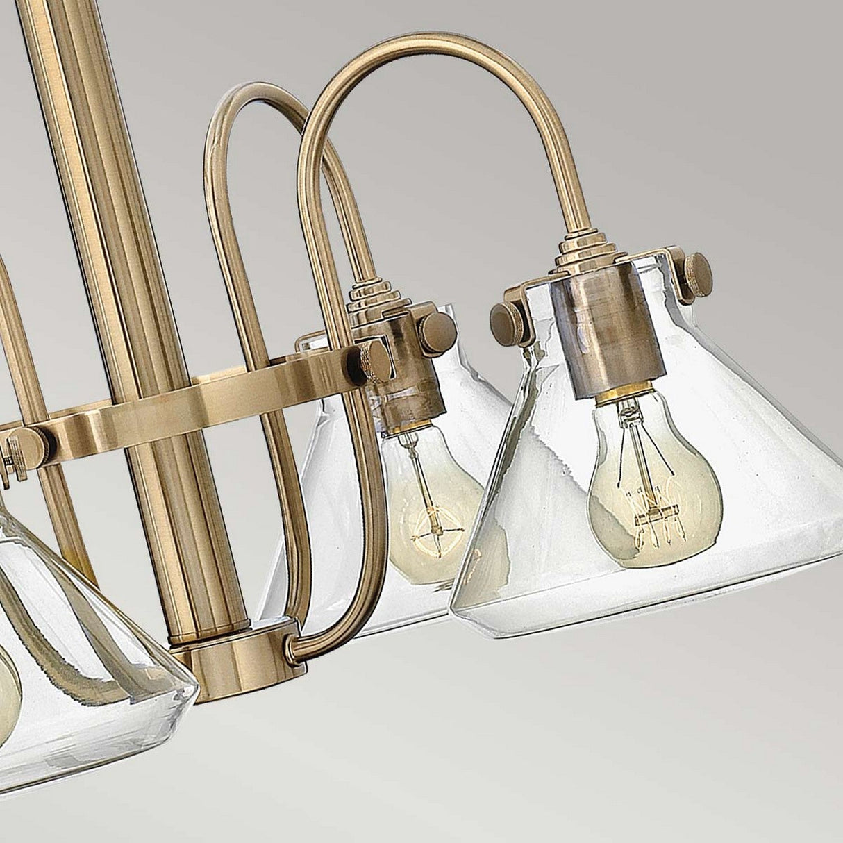 An up-close view of the Congress 4 Light Clear Glass Chandelier reveals its cone-shaped shades that prominently display the light bulbs. The chandelier's brushed caramel finish provides a vintage touch, complementing its sleek modern design against a plain background.