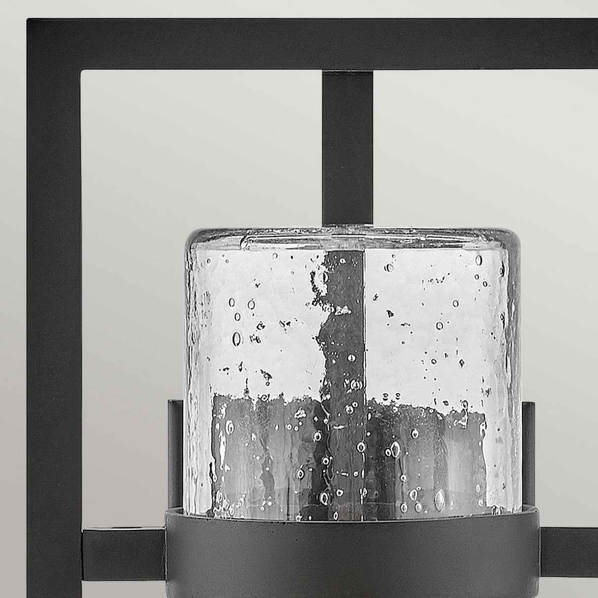 A close-up of the Cordillera 1 Light Outdoor Wall Light showcases its sleek black finish and cylindrical glass shade. The glass, adorned with scattered bubbles, adds texture to the piece. Its modern frame contrasts elegantly against a solid, neutral background.