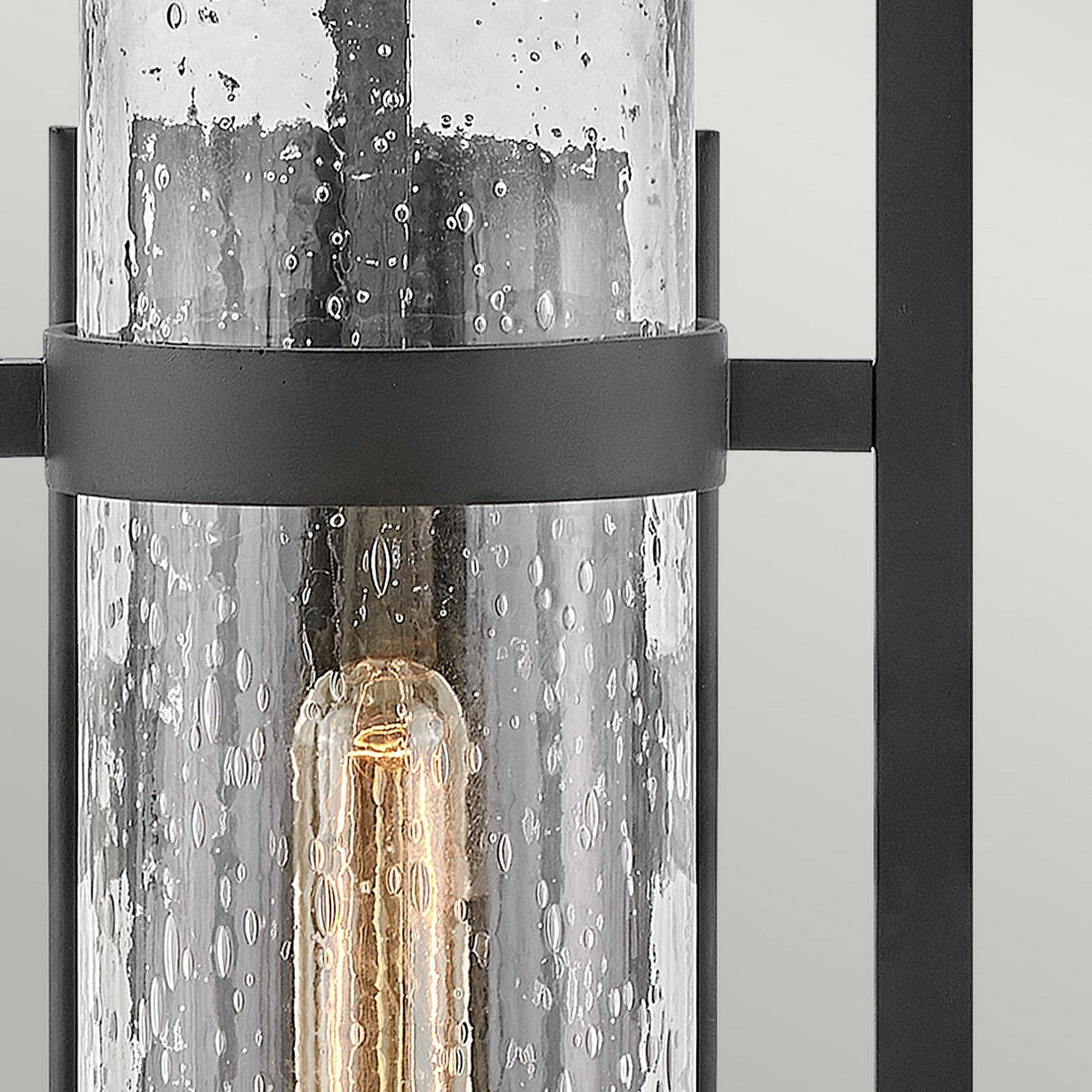 The Cordillera 1 Light Outdoor Wall Light - Black showcases a contemporary design with a modern wall sconce. It features an exposed light bulb enclosed in a clear seeded glass cylinder and includes sleek black metal accents and brackets, making it the perfect choice for outdoor lighting.