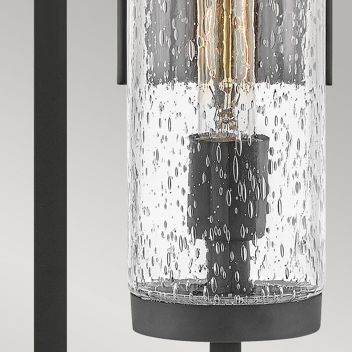The Cordillera 1 Light Outdoor Wall Light - Black features a cylindrical glass fixture with a bubbled texture, encasing vintage-style filament bulbs. It is finished in black and supported by dark metal rods against a light grey background.
