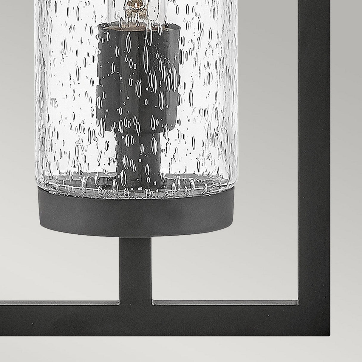 Close-up of the Cordillera 1 Light Outdoor Wall Light - Black, showcasing a modern design with a cylindrical glass cover featuring a bubble pattern. The glass encases a vertical bulb, all set within a sleek black finish frame on a light gray background.
