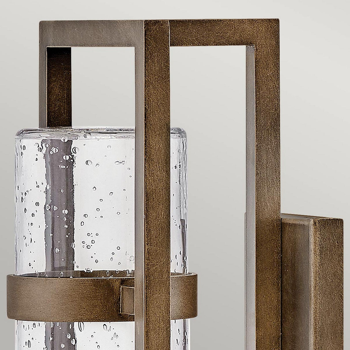 Close-up of the Cordillera 1 Light Outdoor Wall Light in burnished bronze, showcasing its rectangular frame and bubbled-textured cylindrical glass cover. The background emphasizes the sleek, modern design against a simple, light-colored backdrop.