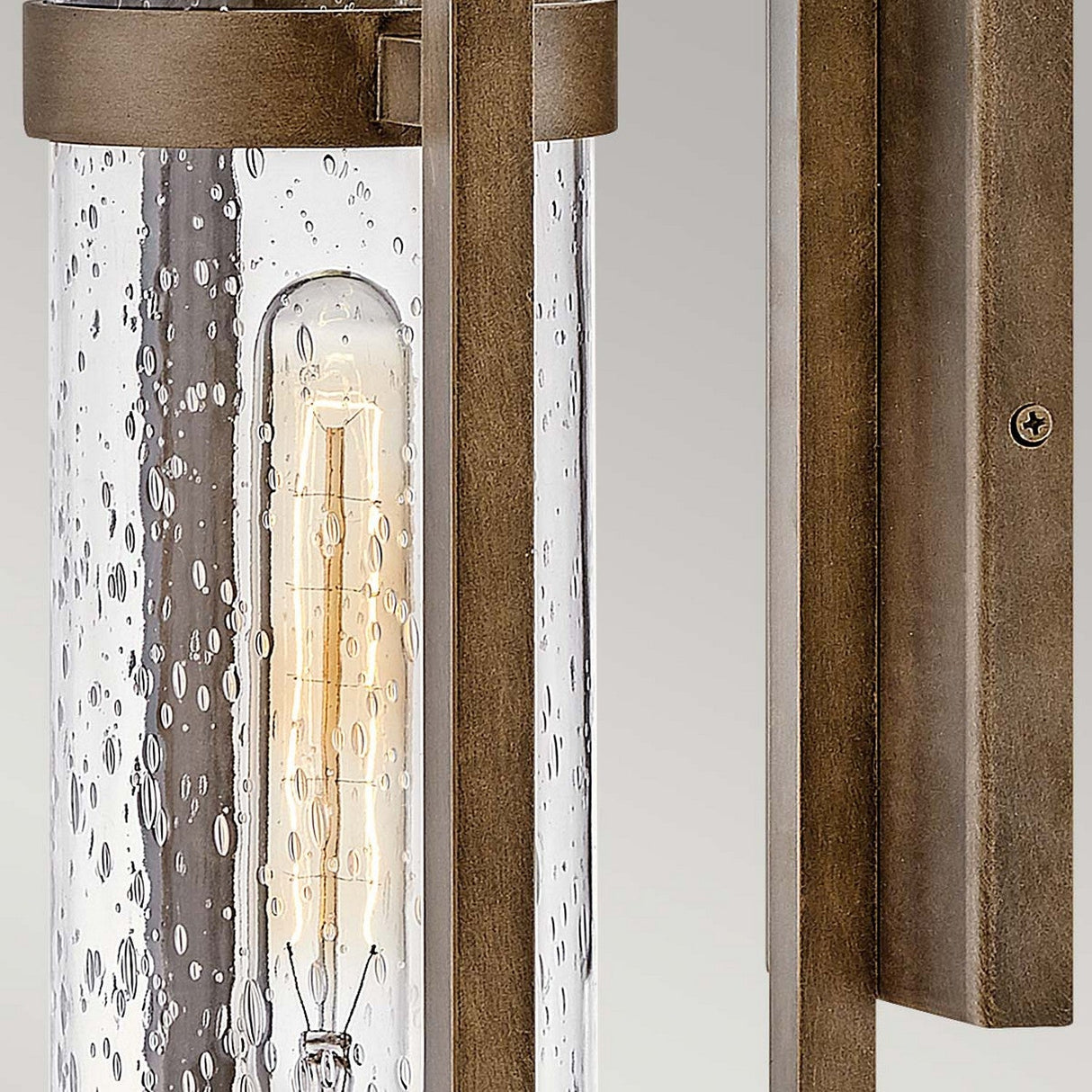 The Cordillera 1 Light Outdoor Wall Light in burnished bronze features a close-up view of its modern design. The cylindrical clear seeded glass shade beautifully displays an exposed vintage-style bulb, all set against a sleek rectangular backplate that merges modern aesthetics with timeless charm.