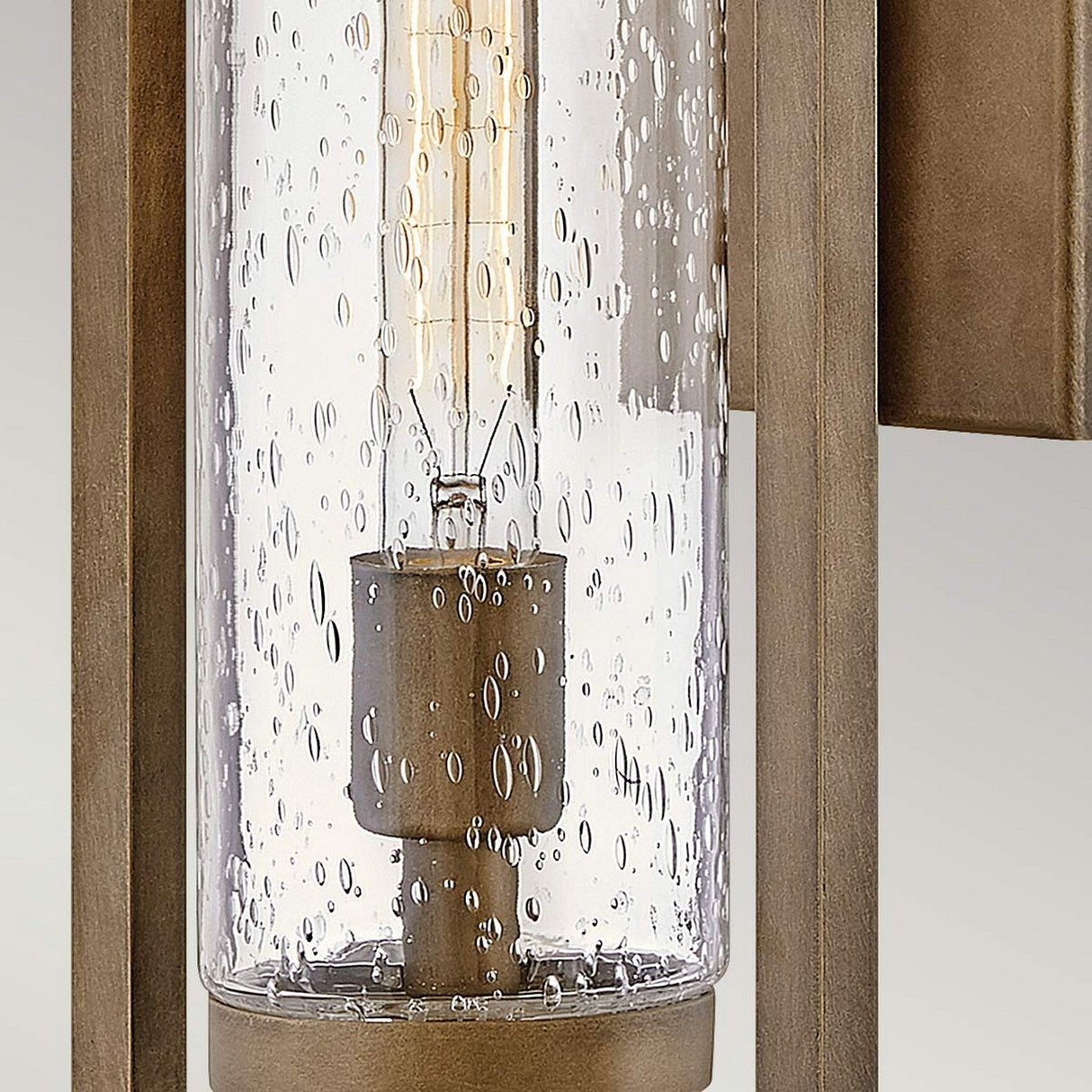 A close-up of the Cordillera 1 Light Outdoor Wall Light showcases its vintage design, with a clear glass cylinder adorned with small water droplet patterns. The fixture is finished in burnished bronze, offering a classic and elegant look while seamlessly integrating modern aesthetics.