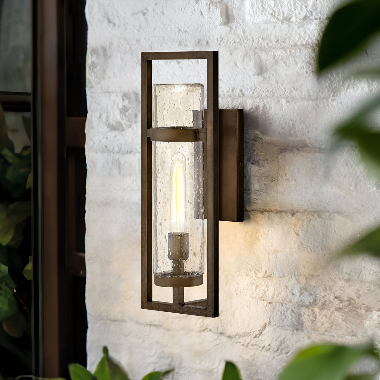 The Cordillera 1 Light Outdoor Wall Light in burnished bronze showcases a sleek design with a textured glass casing. It casts a warm glow against the white brick wall, complemented by nearby green plant leaves, enhancing your exterior decor with its modern aesthetics.
