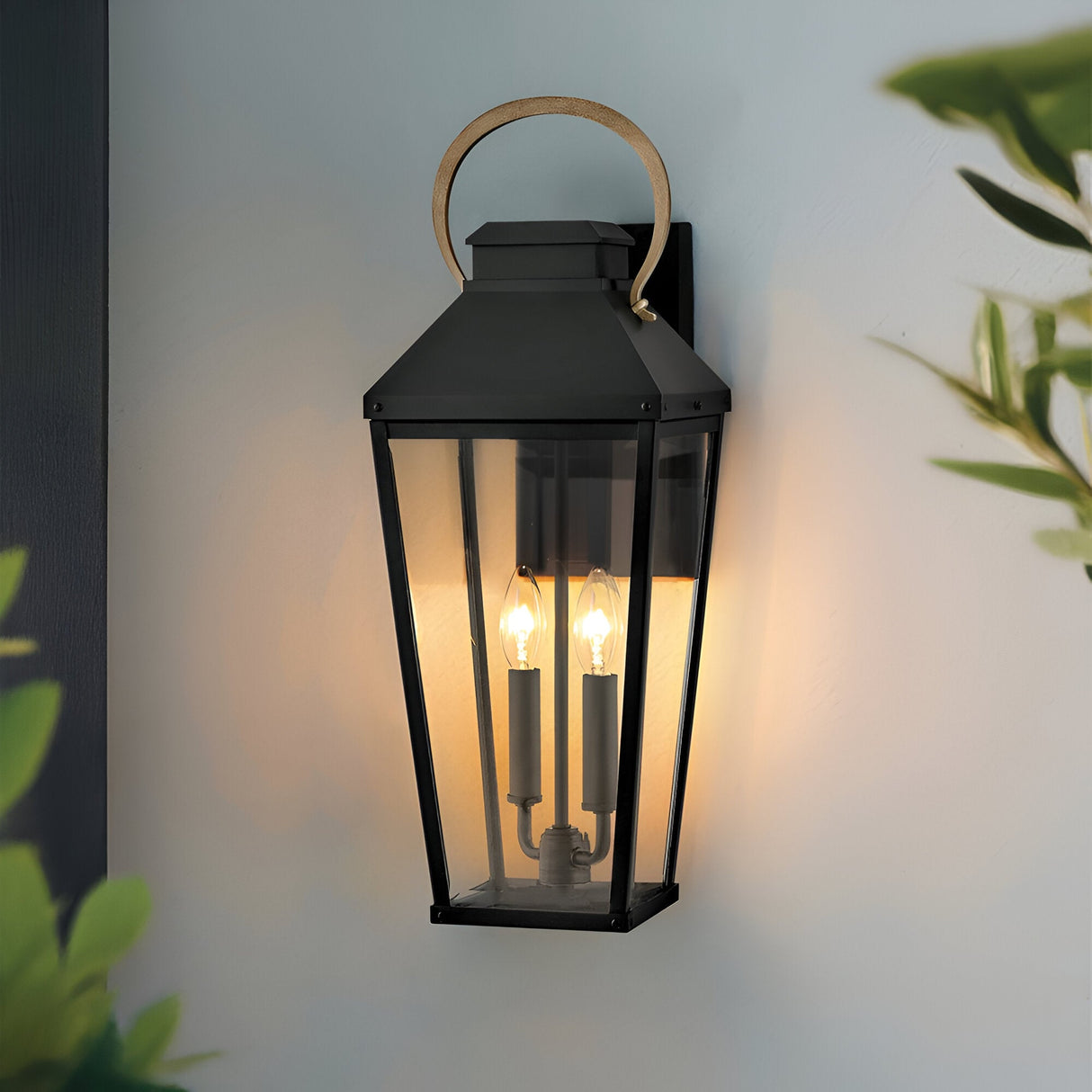 The Dawson 1 Light Outdoor Large Wall Lantern in Black Oxide features glass panels, two candle-style bulbs, and a curved brass handle, providing elegant exterior lighting on light walls amid green foliage.