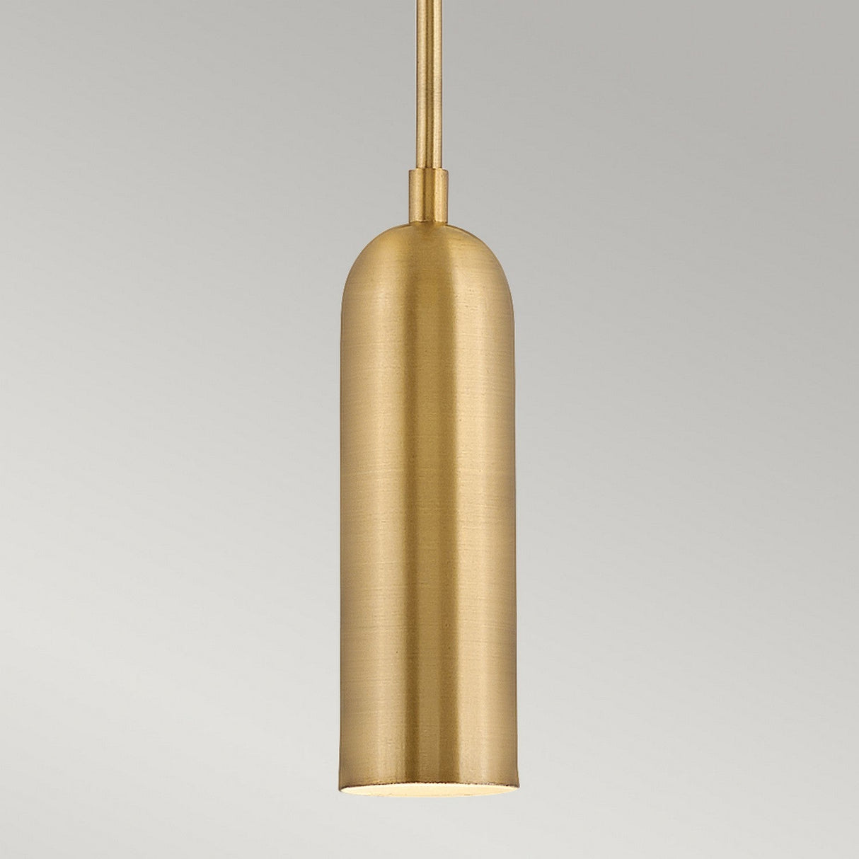The Dax LED Mini Pendant in Heritage Brass, boasting a smooth finish, hangs against a neutral gray backdrop. This pendant offers an energy-efficient lighting solution with its simple, modern design featuring an elongated shape and narrow top.