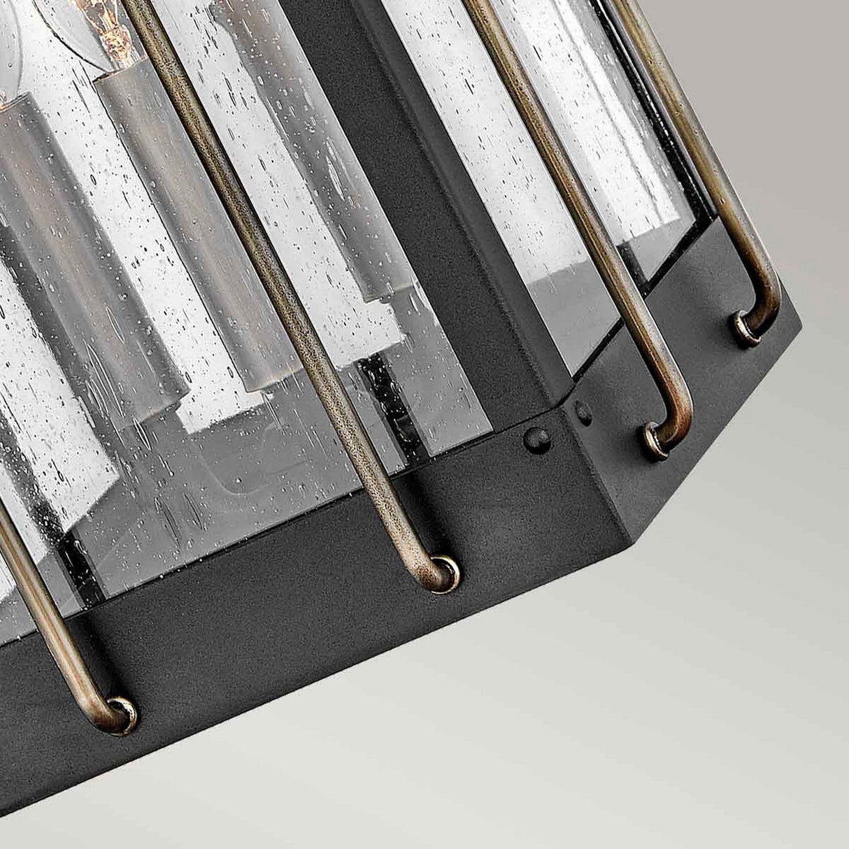 Close-up of the Fitzgerald 3 Light Outdoor Large Wall Lantern, featuring a black hexagonal design with clear, textured glass panels and burnished bronze metal accents that showcase candle-style bulbs inside. This industrial lighting piece is crafted from durable aluminum against a neutral light gray backdrop.