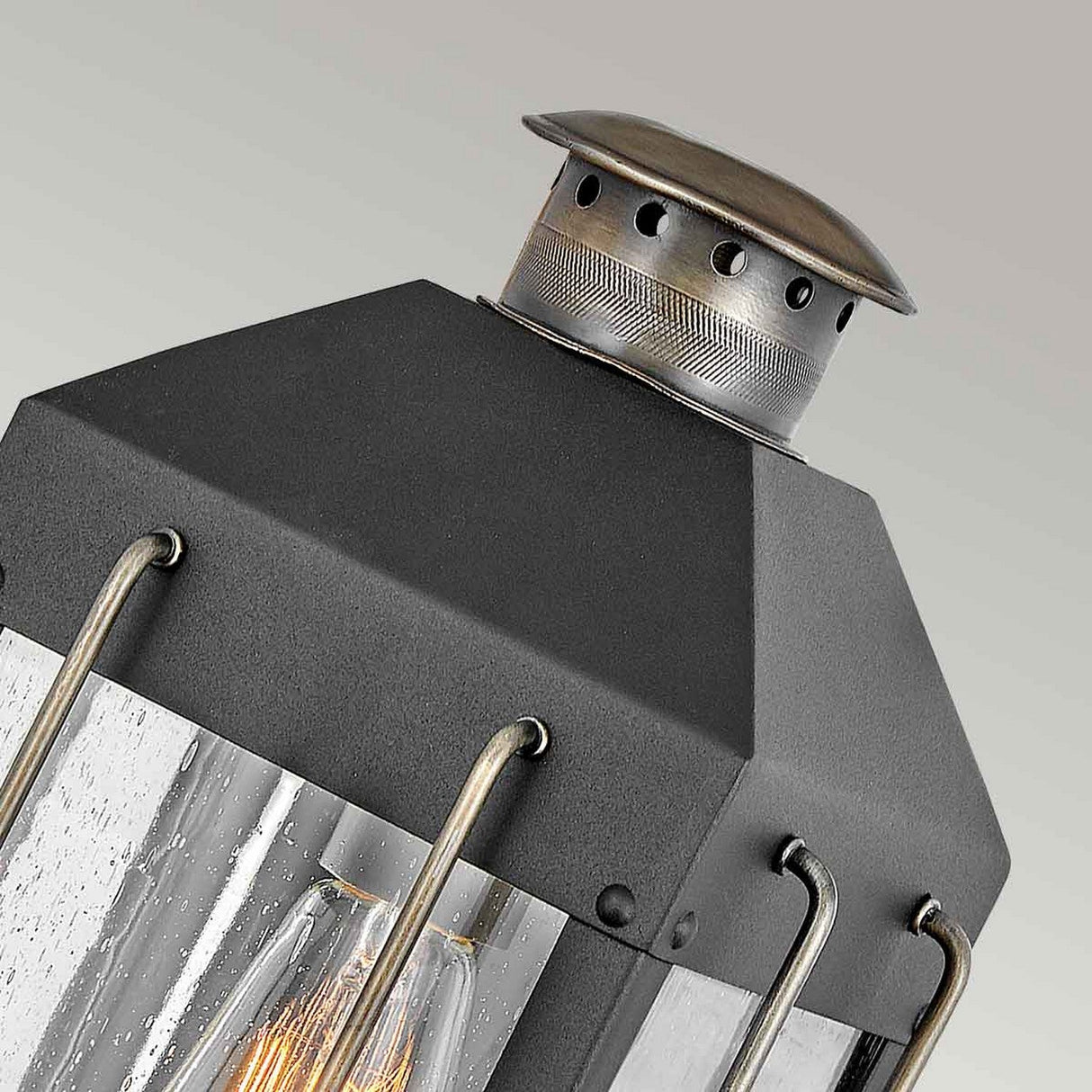 Close-up of the Fitzgerald 1 Light Outdoor Small Wall Lantern, showcasing its vintage-style industrial design. This lantern features a durable aluminum frame with clear textured glass panels. It has a black and burnished bronze metallic top adorned with decorative holes and visible screws, set against a neutral background.