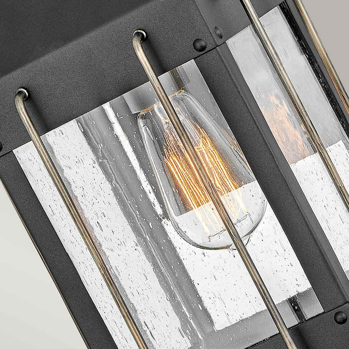 Close-up of the Fitzgerald 1 Light Outdoor Small Wall Lantern in black with burnished bronze, showcasing a vintage-style light bulb glowing warmly inside. The lantern features glass panels with a textured, rain-like pattern and metal bars that enhance its industrial design's rustic and cozy aesthetic. Crafted from durable aluminum for lasting charm.