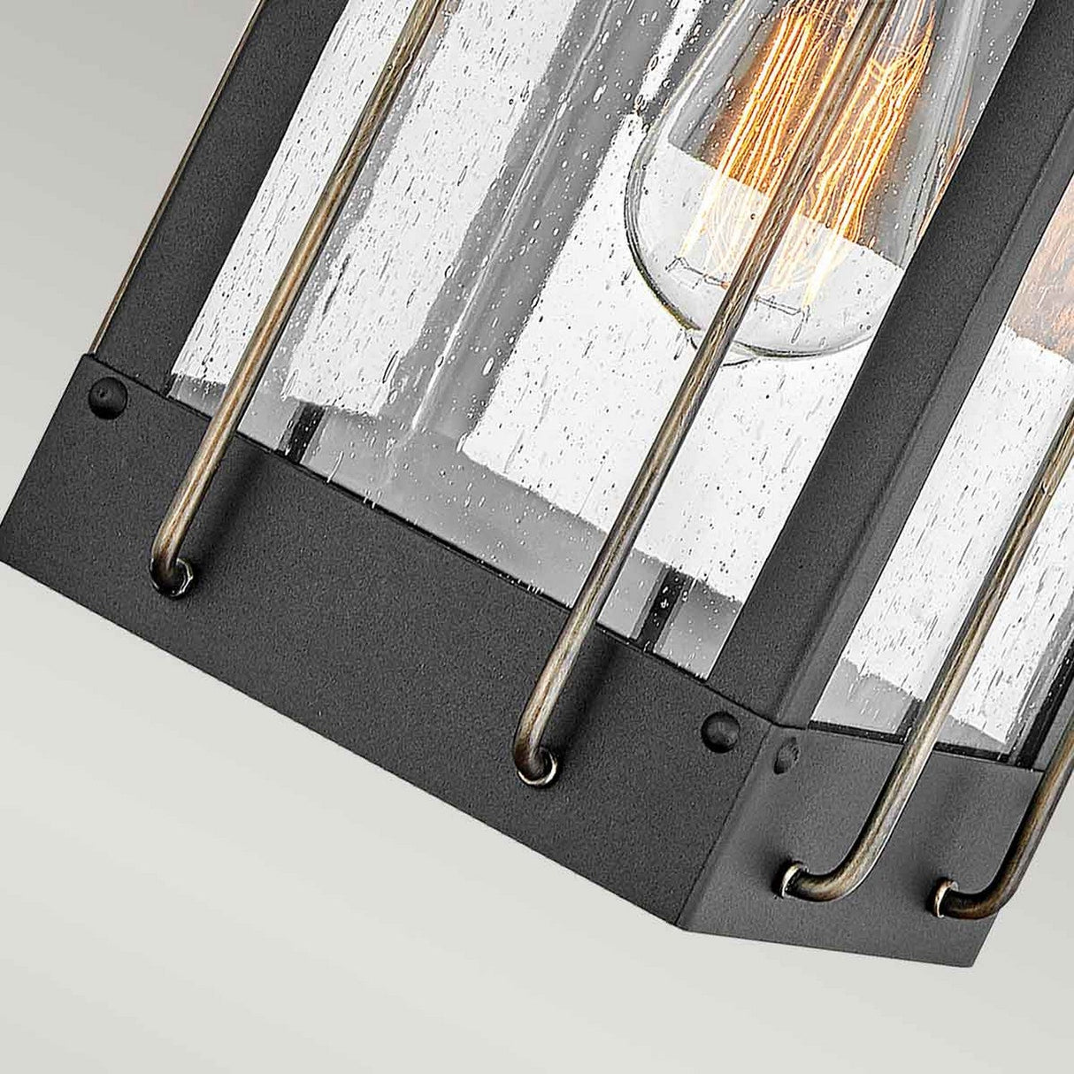 Close-up view of the Fitzgerald 1 Light Outdoor Small Wall Lantern in Black with Burnished Bronze, crafted from durable aluminum and featuring clear glass panels. Inside, an exposed filament bulb emits a warm glow. The sleek, industrial design makes it perfect for contemporary exteriors.