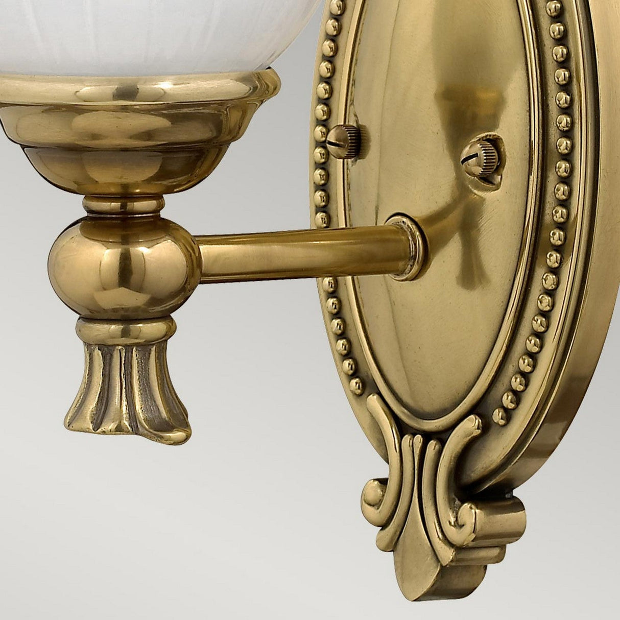 Close-up of the Francoise 1 Light Wall Light in burnished brass, showcasing its ornate decorative mounting plate and vintage-style arm and socket with intricate detailing. The frosted ribbed glass adds texture, set against a soft gray gradient background.