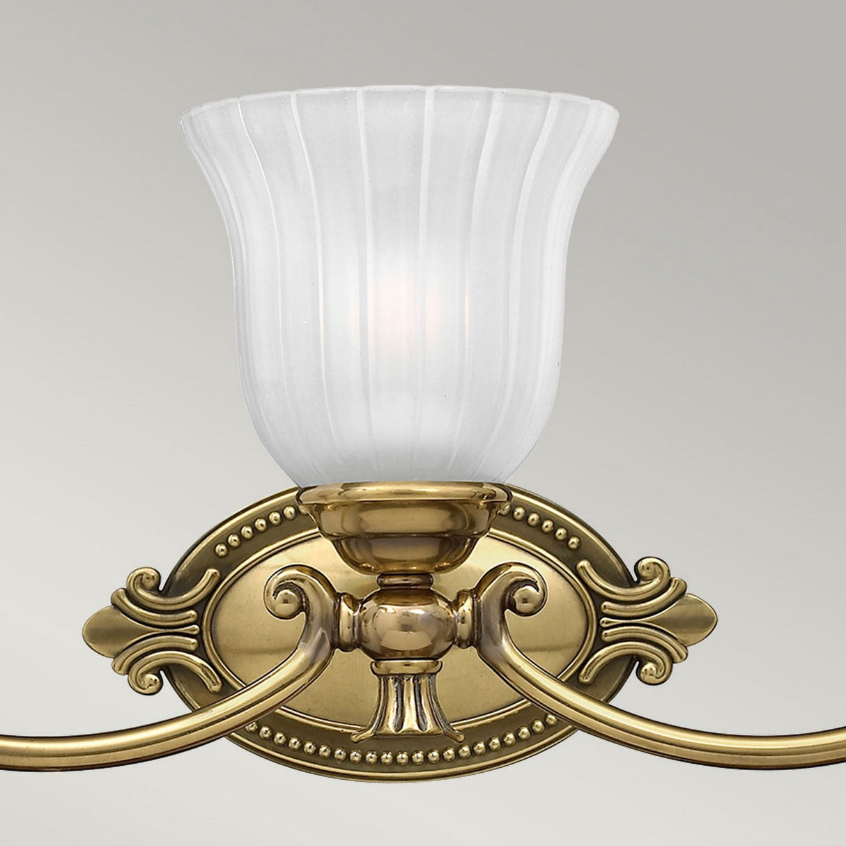 The Francoise 3 Light Wall Light in Burnished Brass features a decorative design with frosted ribbed glass shades and ornate detailing. Its elegant curves and classical motifs exude a vintage charm, making it perfect for mounting against a light gray background.