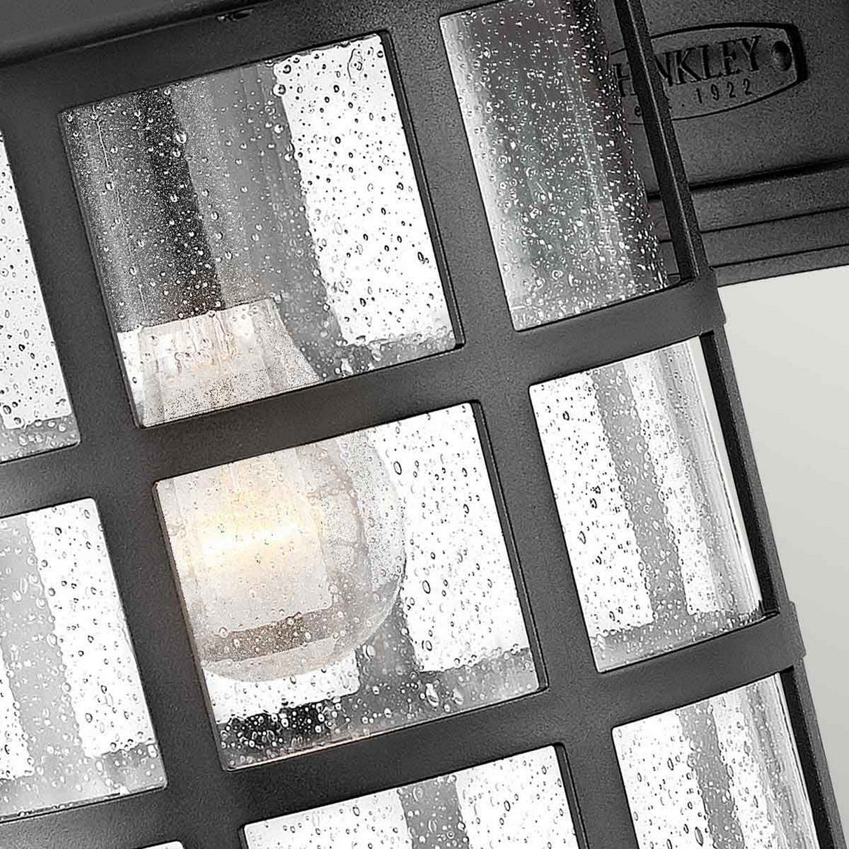 Close-up of the Freeport 1 Light Outdoor Large Wall Lantern - Black, showcasing its weather-resistant design with a grid pattern and rain droplets on the glass. Inside, a glowing light bulb is visible, creating a warm ambiance. The lantern features a sleek black finish and is marked "Since 1922" at the top.