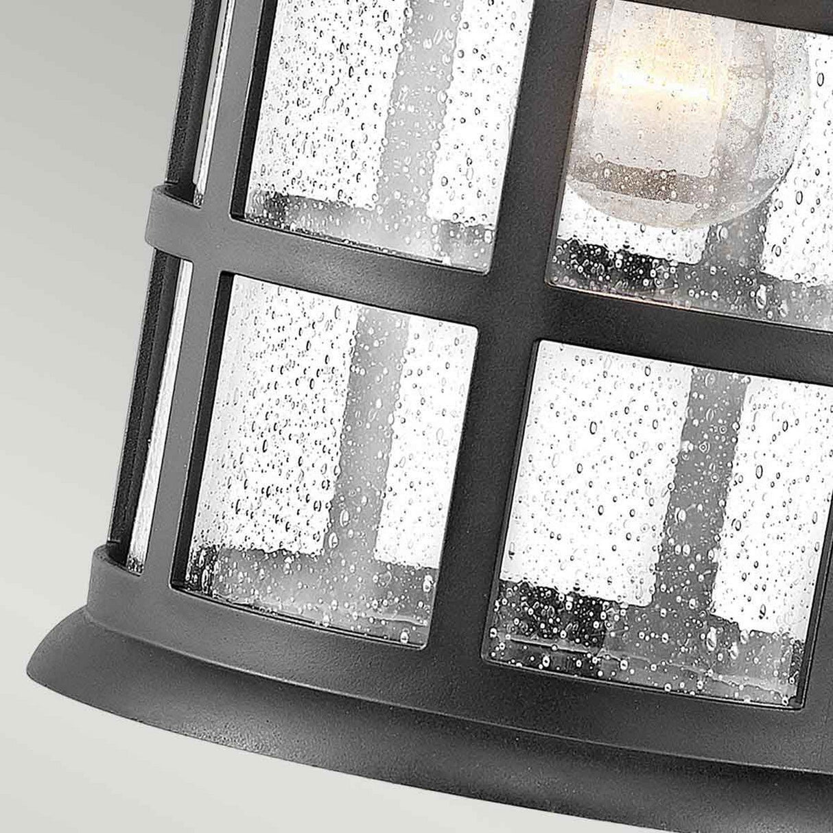 Close-up of the Freeport 1 Light Outdoor Large Wall Lantern - Black, showcasing a grid design with weather-resistant glass panels adorned with water droplet speckles. Inside, a glowing light bulb emits a warm and inviting ambiance.