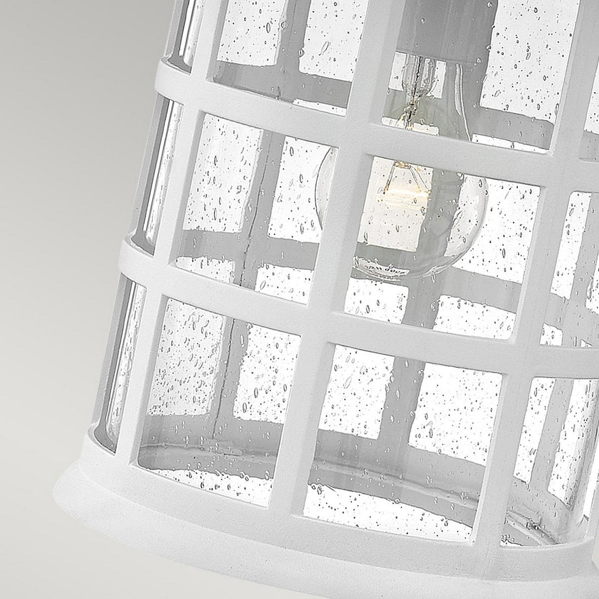 Close-up of the Freeport 1 Light Outdoor Large Wall Lantern in white, featuring clear seeded glass with a grid design that highlights raindrops on the glass surface. A visible light bulb inside underscores its functional performance in wet conditions. Constructed with durable and weather-resistant materials, this lantern promises longevity against the elements. The background is a soft gradient gray.