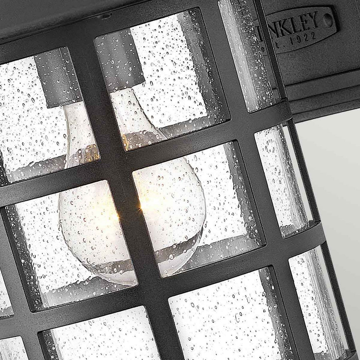 The Freeport 1 Light Outdoor Medium Wall Lantern - Black showcases a detailed view of its vintage-style metal grid design. This weather-resistant lantern is adorned with glass panels shimmering with water droplets, and inside, a glowing bulb creates a cozy ambiance.