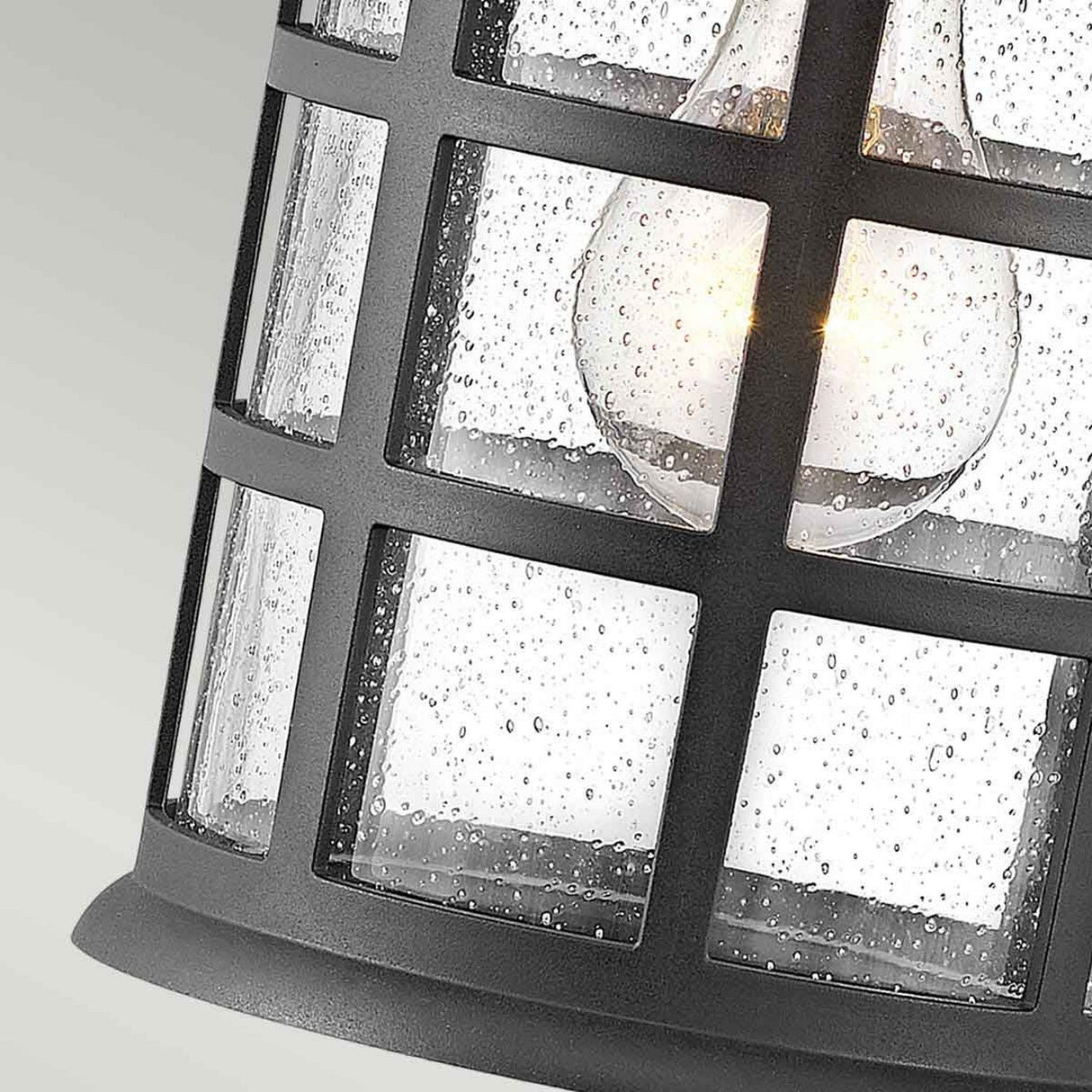 Close-up of the Freeport 1 Light Outdoor Medium Wall Lantern in black, showcasing its curved design and clear bubbled glass enclosure. A glowing light bulb inside emits a warm glow. This weather-resistant lantern stands out against a soft gradient background of light green.