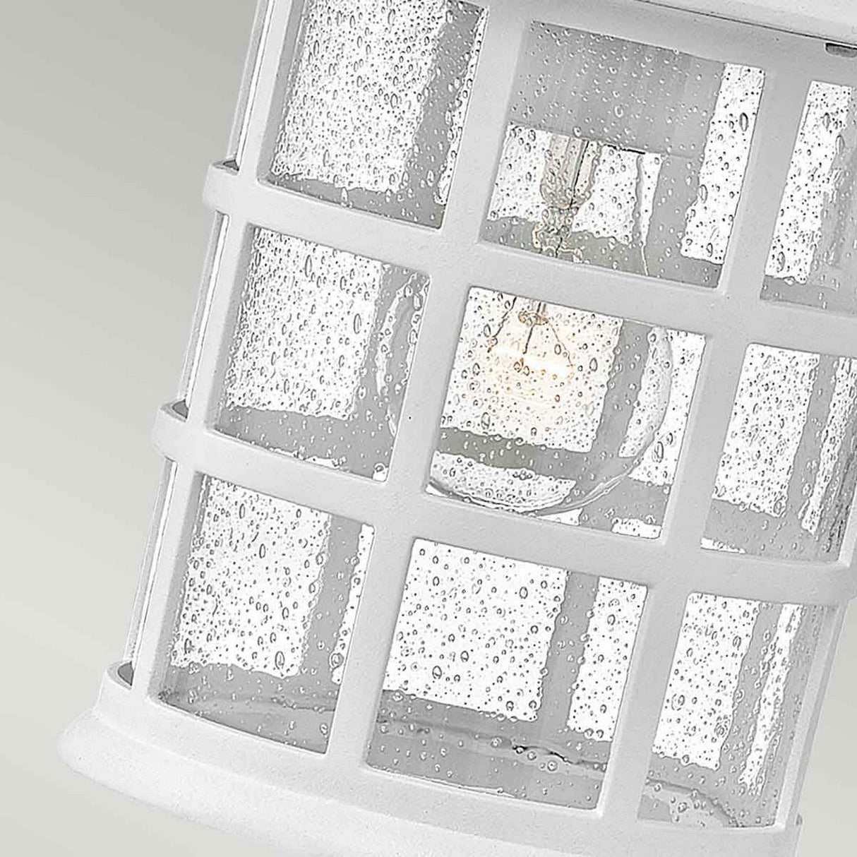 A close-up of the Freeport 1 Light Outdoor Medium Wall Lantern in white showcases a grid-patterned cylinder and droplets on its clear seeded glass shade. Inside, the light bulb emits a soft glow, enhancing its weather-resistant design against a light green gradient backdrop.