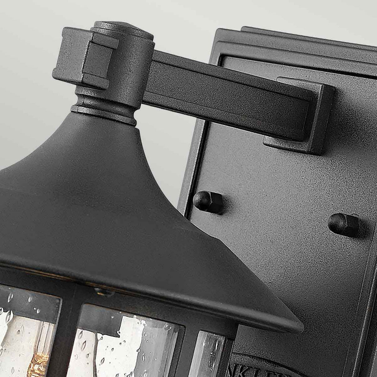 A close-up of the Freeport 1 Light Outdoor Small Wall Lantern in black showcases its weather-resistant, lantern-style design. The fixture boasts a sturdy metal frame and a glass enclosure adorned with decorative rain droplets, all set against a neutral gray wall.