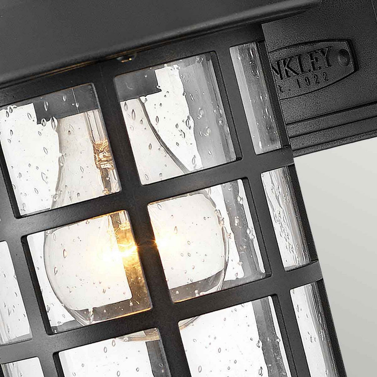 Close-up of the Freeport 1 Light Outdoor Small Wall Lantern - Black, featuring a weather-resistant finish and a glowing light bulb inside. The glass panes are speckled with raindrops, and a small plaque on the lantern is inscribed with Kichler 1992 in raised letters.