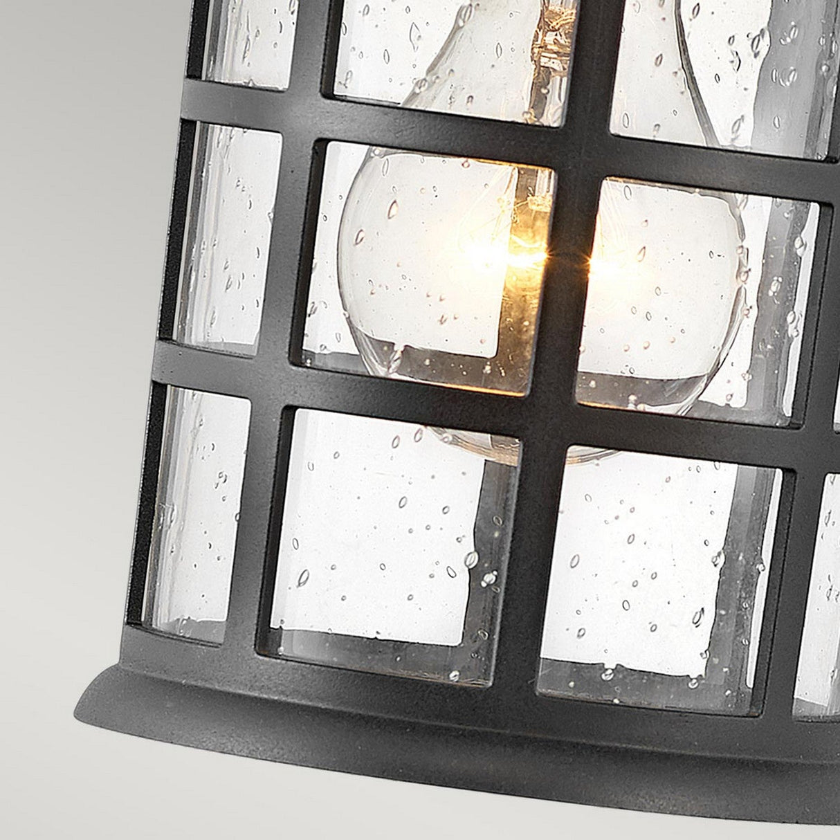 A close-up of the Freeport 1 Light Outdoor Small Wall Lantern in black showcases its weather-resistant design with a metal grid. The bulb inside emits a warm glow, while the glass covering is speckled with water droplets, suggesting recent rain or high humidity. The background is kept simple and neutral.