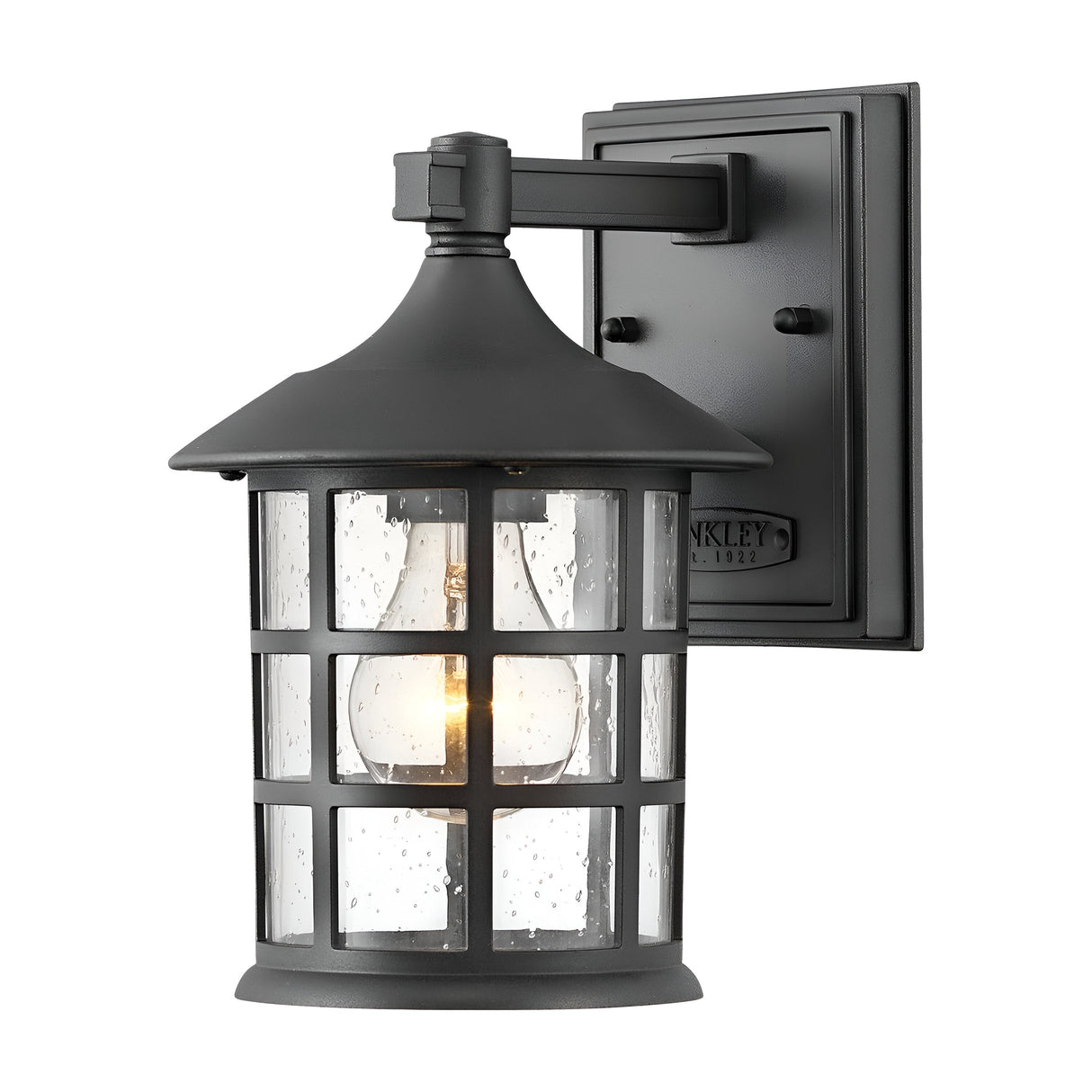 The Freeport 1 Light Outdoor Small Wall Lantern - Black showcases a classic black finish with a domed top and clear glass panels that reveal a glowing light bulb. This weather-resistant fixture is mounted on a rectangular base with an ornate frame design, guaranteeing resilience against the elements.
