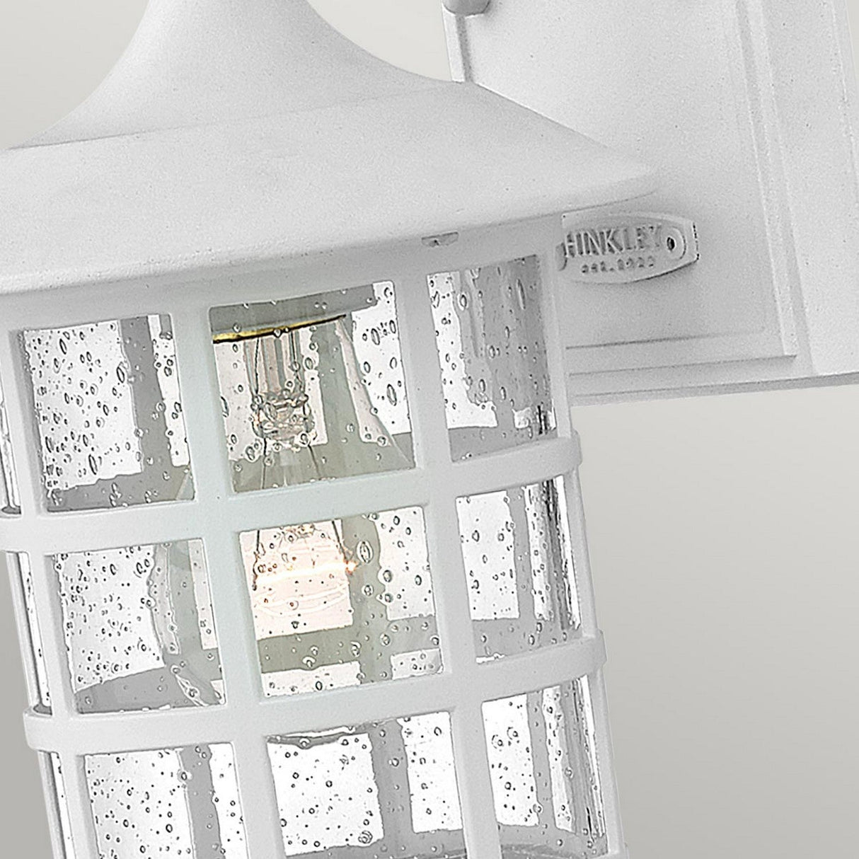Close-up of the Freeport 1 Light Outdoor Small Wall Lantern in white, showcasing its IP44-rated design with grid-patterned, raindrop-textured glass. The visible light bulb inside provides illumination, and a small metal nameplate adorns this weather-resistant fixture.