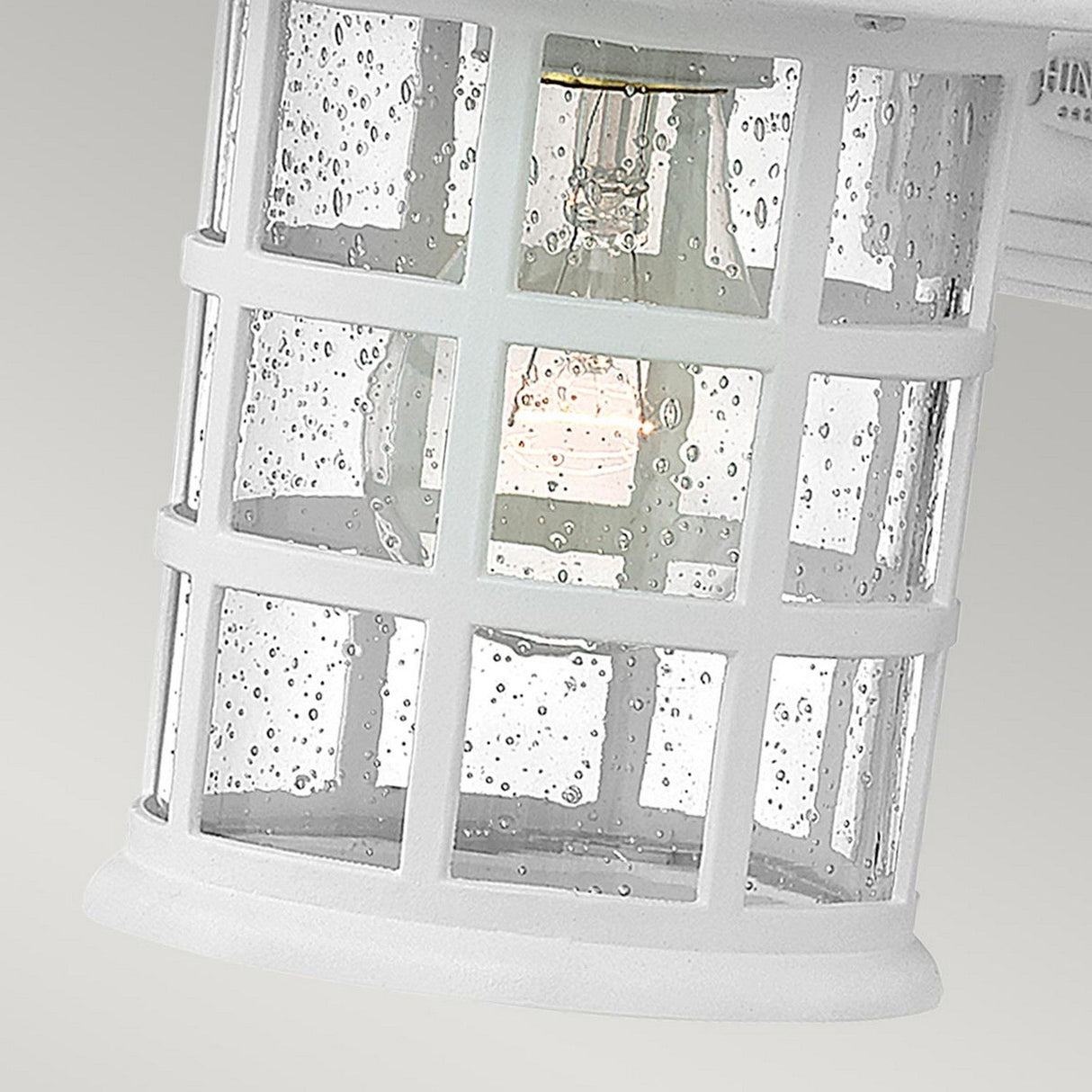 Close-up of the Freeport 1 Light Outdoor Small Wall Lantern in white, showcasing its grid design with a clear glass covering adorned with water droplets. A visible bulb emits a soft glow, accentuating the droplets. The IP44 rated lantern is weather-resistant and set against a muted gray background.