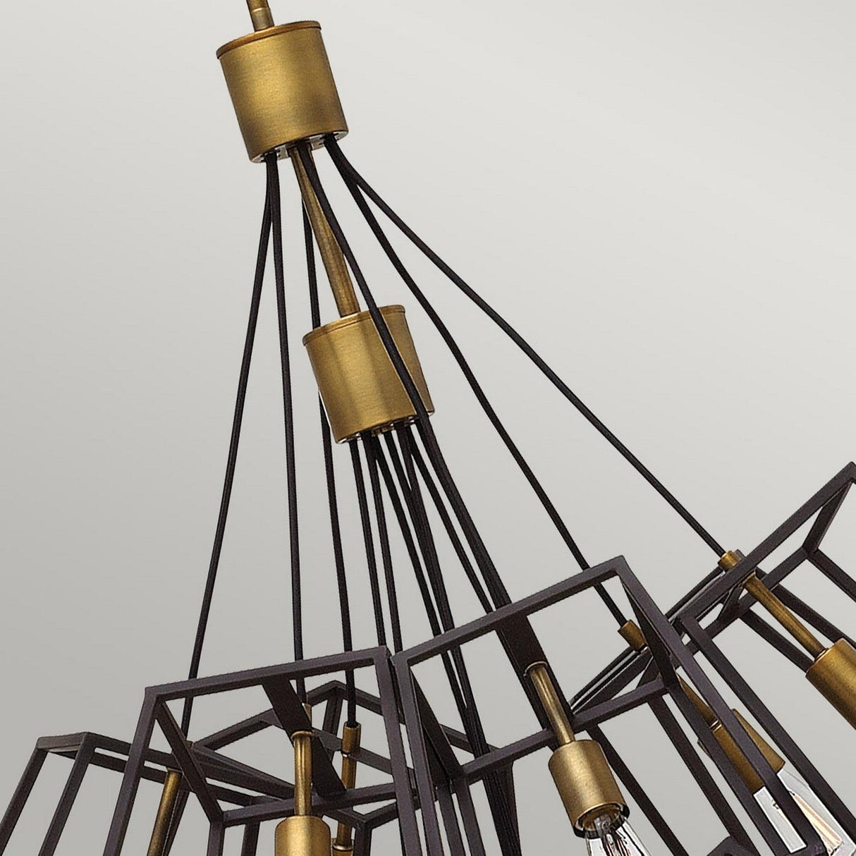 A contemporary chandelier that combines a vintage industrial aesthetic, the Fulton 13 Light Large Chandelier - Bronze & Brass features cube-shaped frames arranged around a central axis, suspended by cables connected to a cylindrical fixture. The entire design is set against a plain, light grey background.