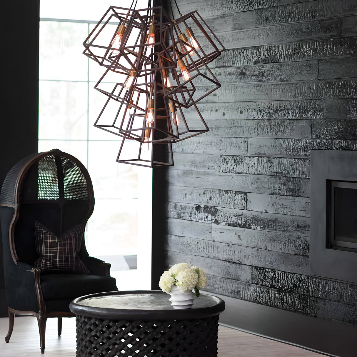 A modern living room with dark wood-paneled walls and a vintage industrial twist, featuring the Fulton 13 Light Large Chandelier in Bronze & Brass. A unique black chair with a plaid cushion complements the round black table adorned with a white floral arrangement, while a large window floods the space with natural light.