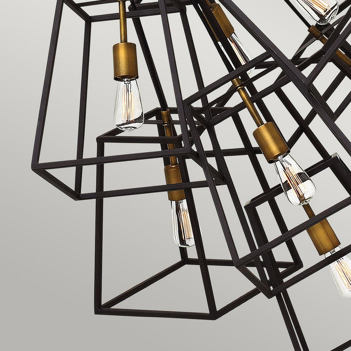 The Fulton 7 Light Chandelier - Bronze & Brass exemplifies minimalist design with its black metal frame featuring exposed Edison-style bulbs and exquisite brass accents, all elegantly contrasted against a plain gray background.