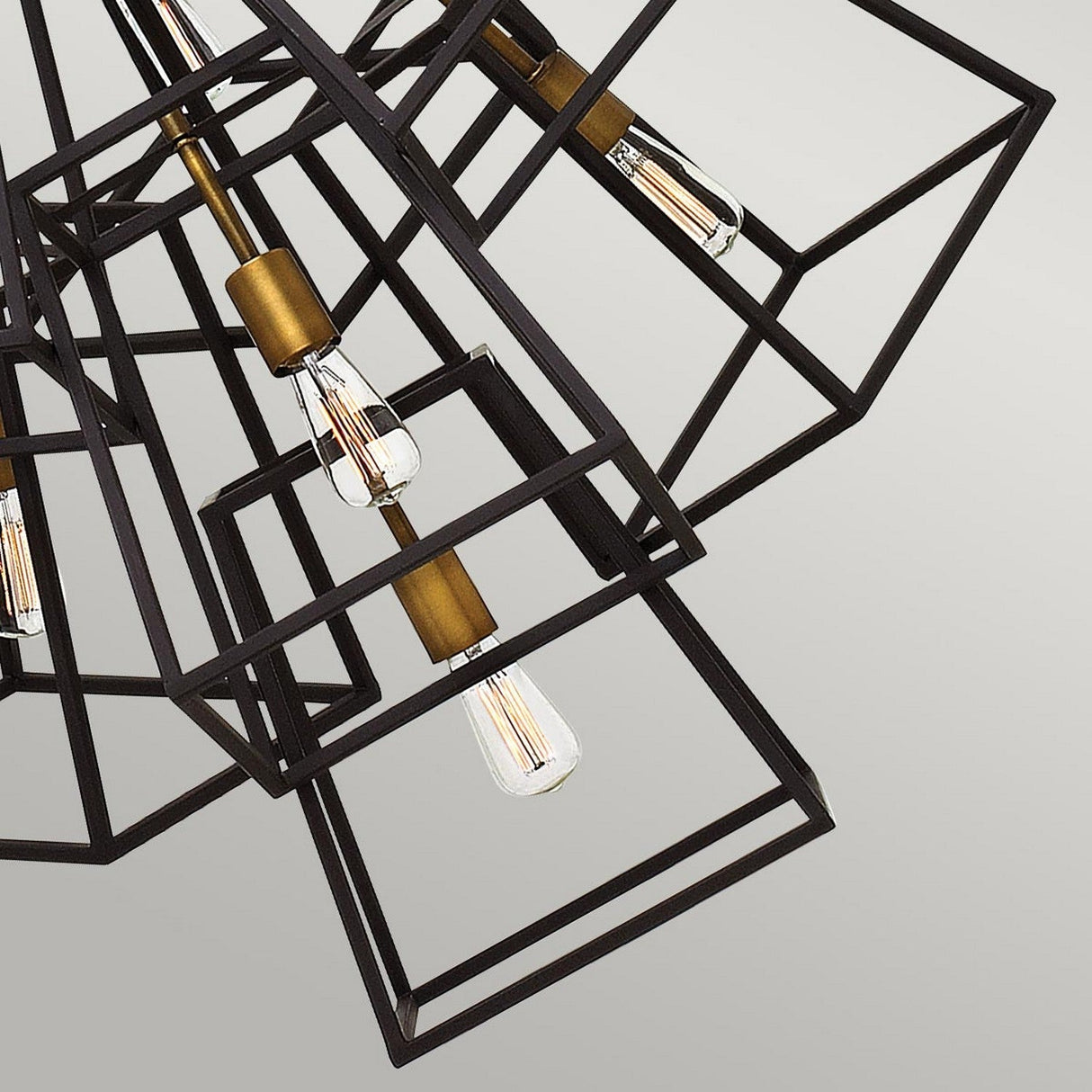 The Fulton 7 Light Chandelier - Bronze & Brass showcases a geometric black metal frame with exposed vintage-style Edison bulbs and bronze fittings, creating a minimalist aesthetic against a light gray background.