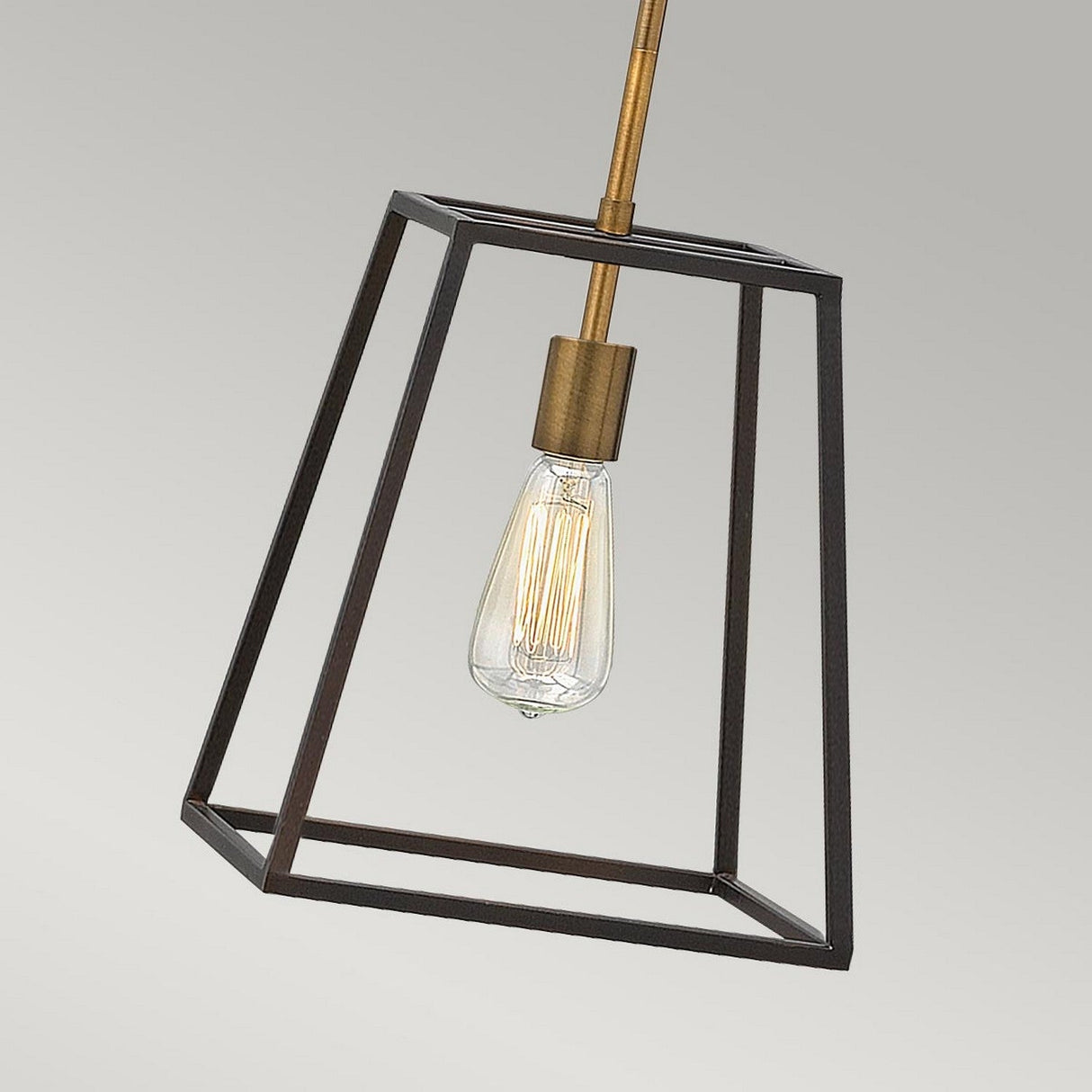 The Fulton 1 Light Pendant in Bronze & Brass showcases a geometric metal frame that encases an Edison bulb, exuding a vintage industrial style. Its minimalist design with a bronze frame and brass accents contrasts beautifully against a plain, light gray background.