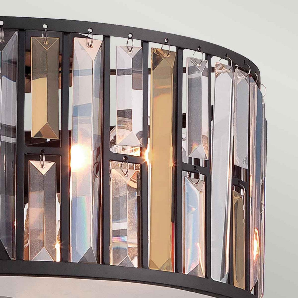A close-up of the Gemma 3 Light Flush in Vintage Bronze showcases its ornate design, featuring vertical rectangular glass pendants that elegantly reflect light. The circular metal frame with a vintage bronze finish complements its modern and sophisticated look against a light background.