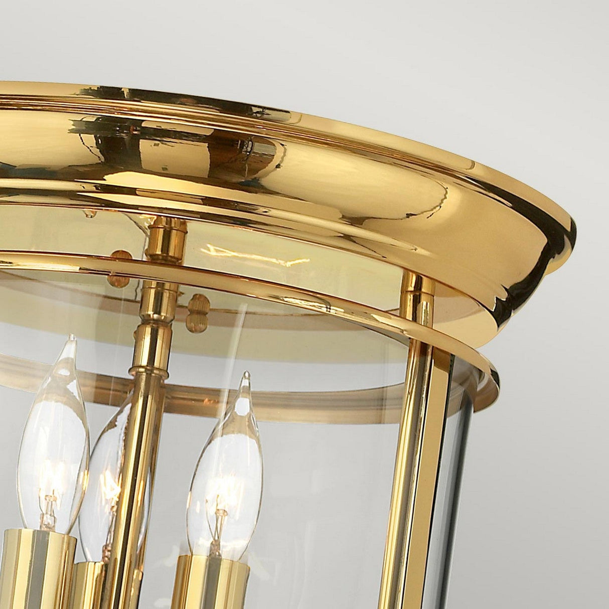 Close-up of the Gentry 3 Light Flush - Polished Brass ceiling light fixture with a clear glass enclosure, featuring three candle-shaped bulbs inside. This traditional style piece adds elegance and charm against a plain white background.