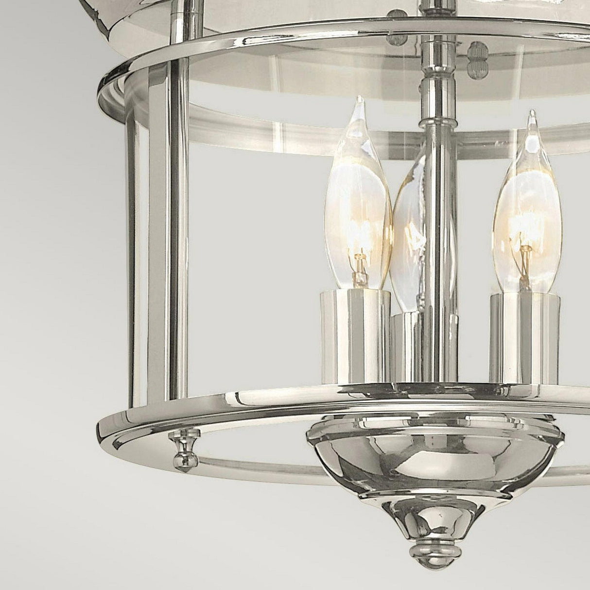 A close-up of the Gentry 3 Light Flush in polished nickel highlights its cylindrical glass shade, within which two candle-shaped bulbs emit a warm glow, offering a nod to traditional design elements. This modern chandelier contrasts beautifully against the simple light gray backdrop.
