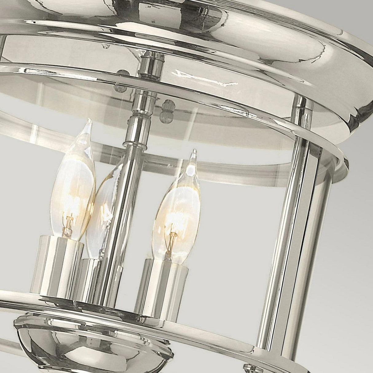 Close-up of the Gentry 3 Light Flush - Polished Nickel, a ceiling light that combines modern and traditional design elements with three candle-style bulbs encased in a cylindrical frame, set against a neutral background.