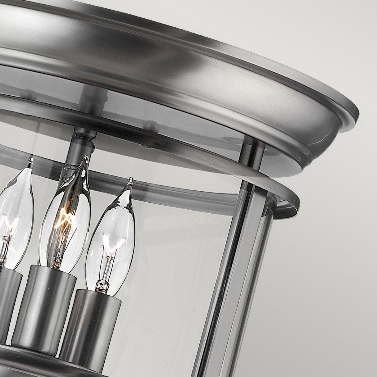 A close-up of the Gentry 3 Light Flush - Silver reveals its modern, brushed metallic charm. Featuring three candle-shaped bulbs enclosed in clear glass, its sleek design includes a circular top and a silver finish, epitomizing contemporary elegance.
