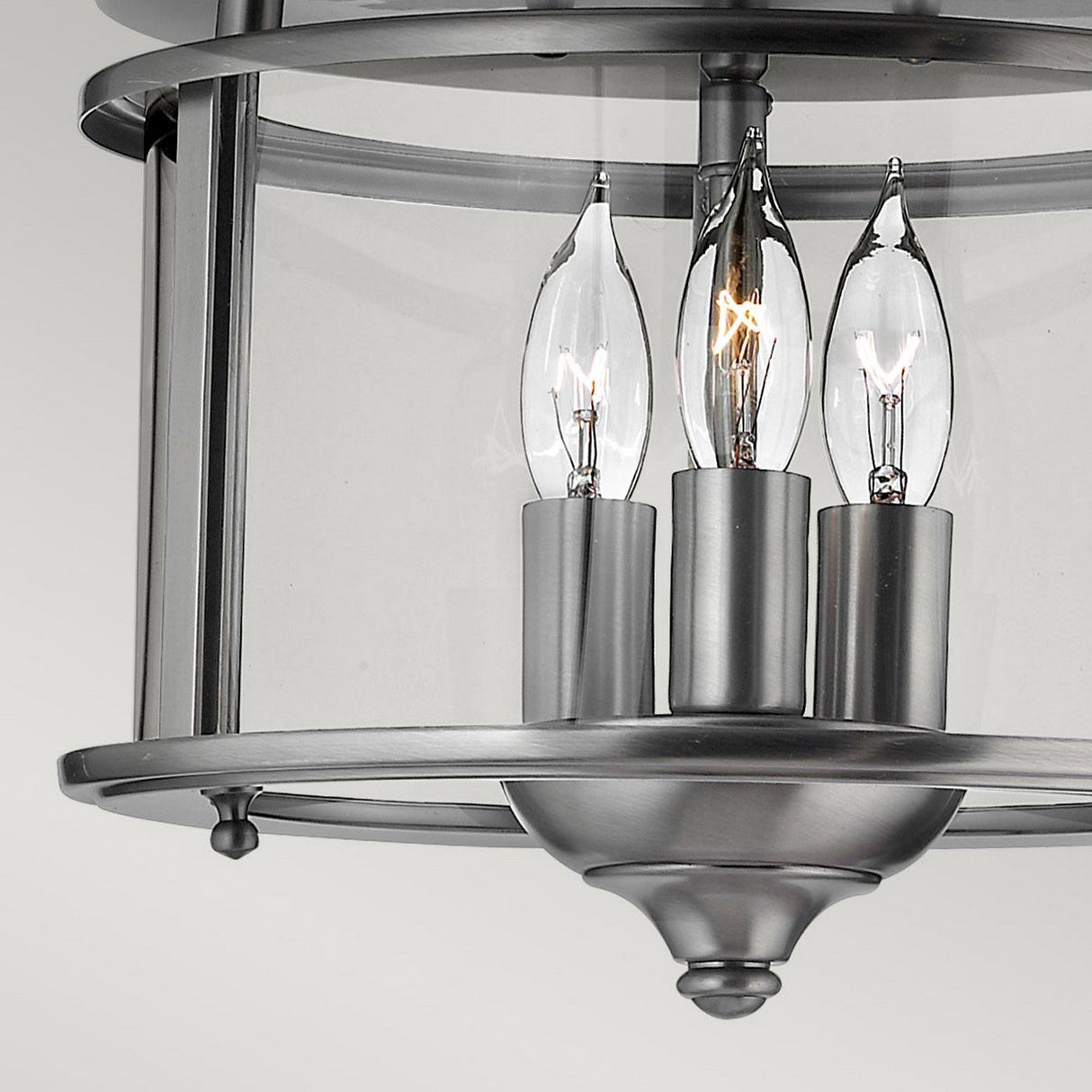 The contemporary ceiling light, Gentry 3 Light Flush - Silver, showcases three candle-shaped bulbs enclosed in a cylindrical clear glass cover. It features a sleek metallic finish and a modern design that complements any decor.