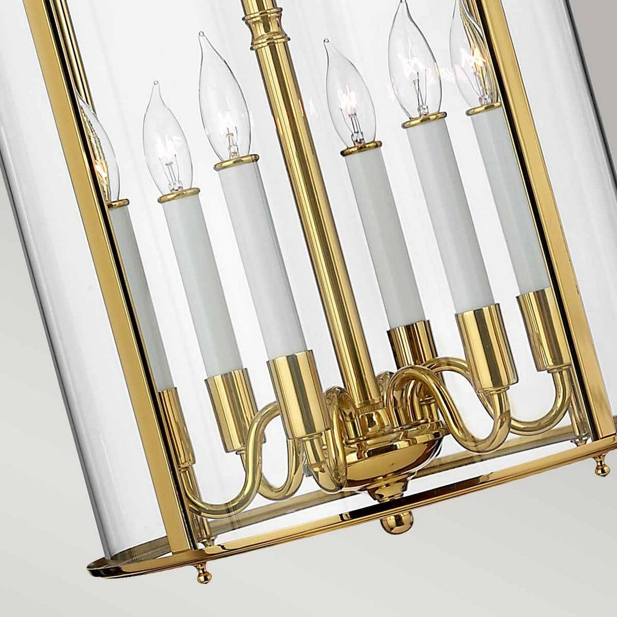 The Gentry 6 Light Large Pendant in polished brass features a traditional design, showcasing tall, candle-shaped lights within clear bent glass enclosures. It is adorned with six flame-like bulbs set against a softly blurred background.
