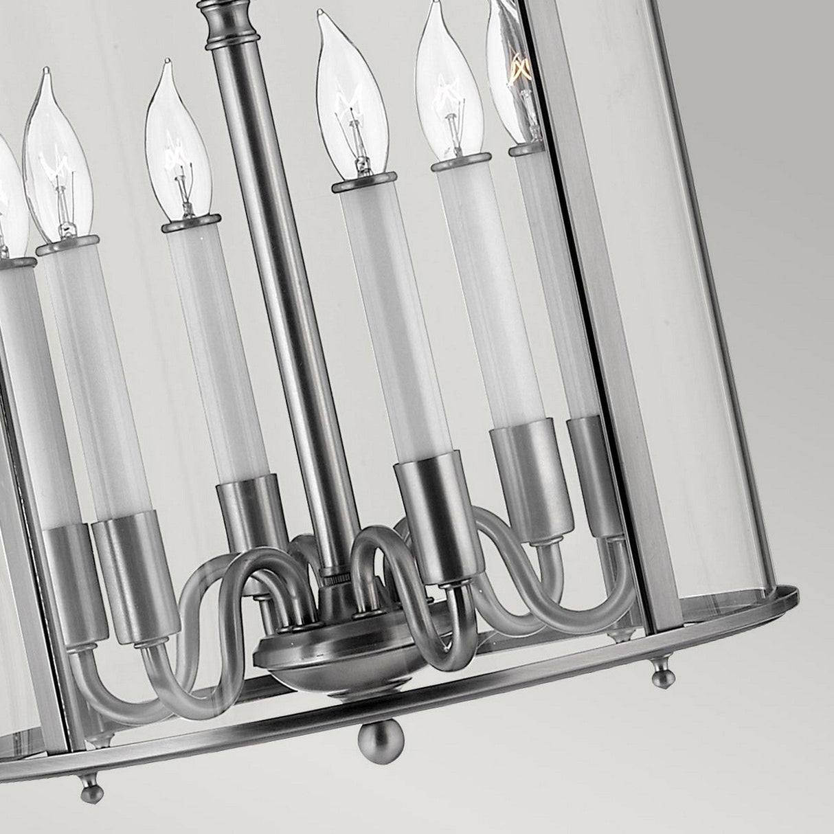 A close-up of the Gentry 6 Light Large Pendant in Silver showcases a sleek, contemporary lighting fixture featuring a cylindrical chandelier design with six candle-like bulb holders. Each holder cradles a clear bulb within a metallic frame with silver undertones, surrounded by a transparent glass cover that enhances its modern appeal.